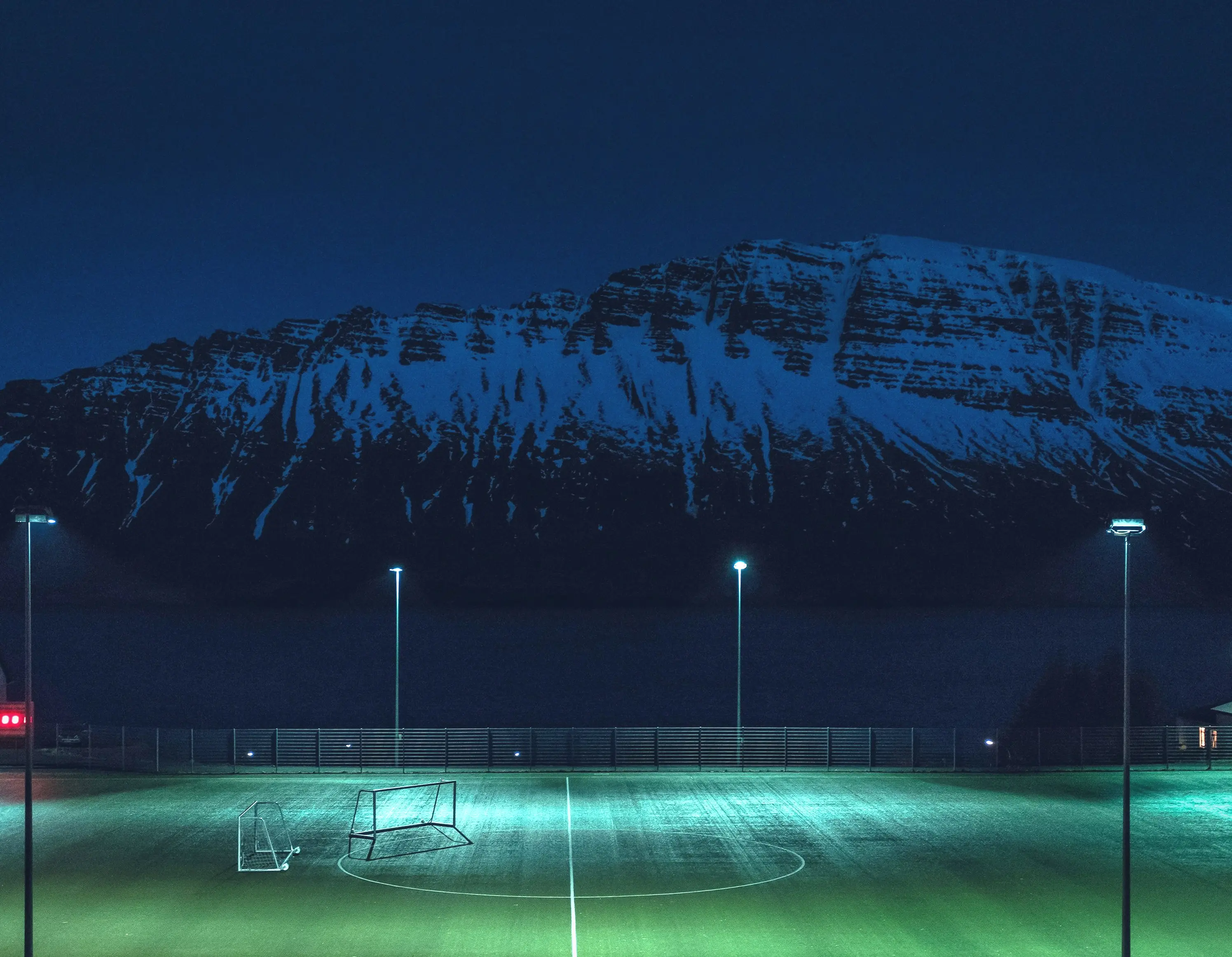 An empty floodlit pitch at dusk beneath a snow-covered mountain